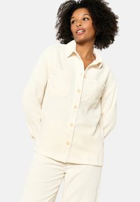 Cream corduroy button-up shirt with two front pockets and a relaxed fit, featuring a collar and ribbed texture. Paired with matching pants.