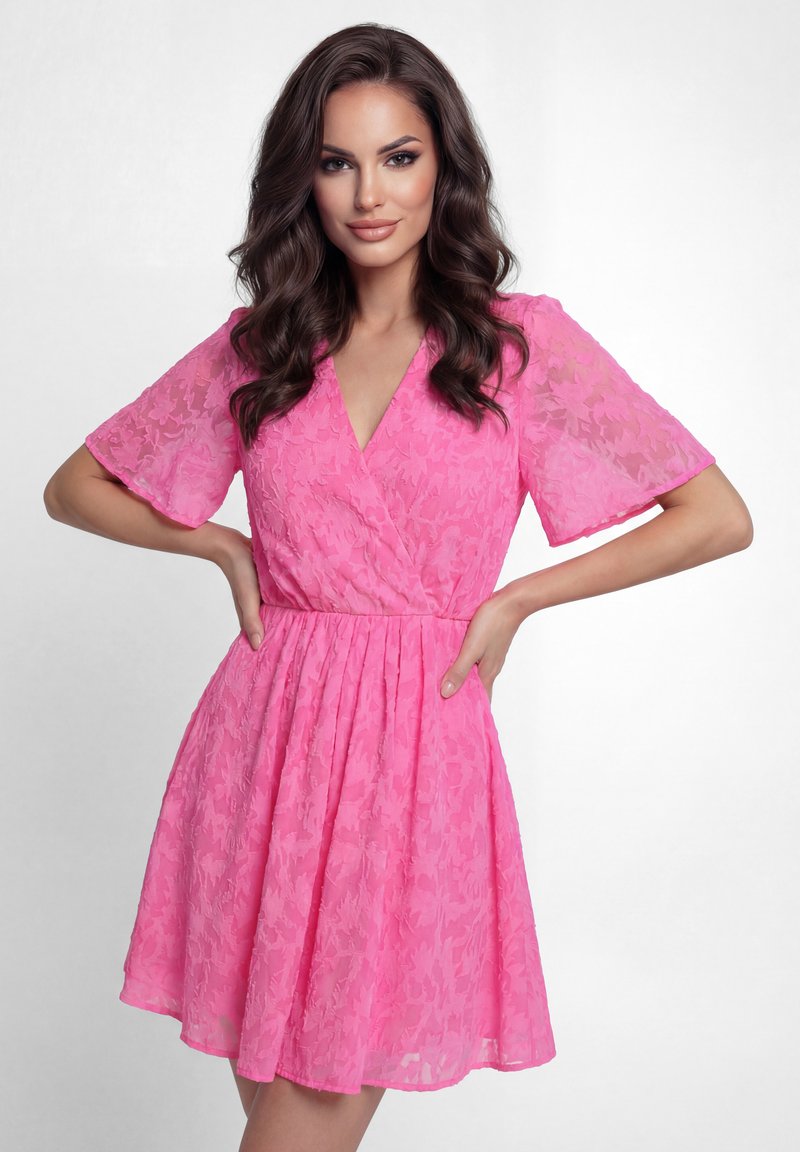 Vrouw met lang bruin haar, gekleed in een roze bloemenjurk met korte mouwen, staat met haar handen op haar heupen tegen een witte achtergrond.