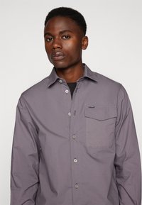 Camisa gris de botones hecha de tela ligera, que cuenta con un bolsillo en el pecho, cuello estándar y una textura suave.