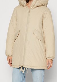 Chaqueta acolchada beige con cierre frontal, grandes bolsillos laterales y capucha. Presenta puños elásticos y un dobladillo ajustable con cordón.