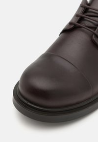 Scarpa in pelle marrone scuro con punta arrotondata, finitura liscia e suola in gomma nera. I lacci passano attraverso occhielli tonalizzati.