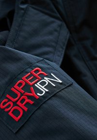 Giacca in tessuto blu navy con un design testurizzato. Presenta una toppa con "SUPERDRY" in rosso e "JPN" in bianco, con dettagli ricamati.