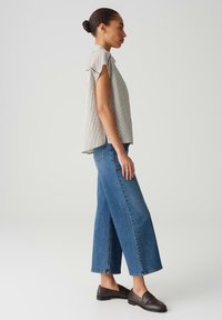 Top a righe a maniche corte in bianco e nero, abbinato a jeans in denim blu a gamba larga. Modella indossa scarpe slip-on scure, vista laterale.