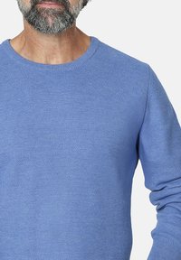 Hombre barbudo que lleva un suéter de manga larga de textura azul claro con un cuello redondo, recortado en los hombros y a la altura del torso.