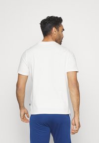 Hvid bomulds t-shirt med rund hals og korte ærmer, vist fra ryggen. Bæres med blå bukser, der har lige kant og afslappet pasform.