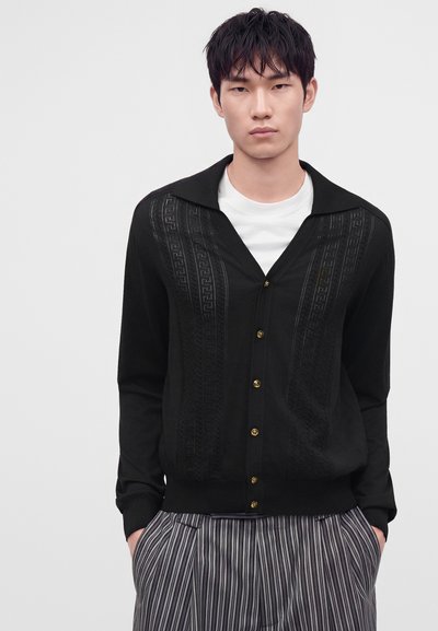 Giovane uomo con capelli corti e neri indossa un cardigan nero abbottonato sopra a una camicia bianca e pantaloni grigi a righe, in piedi davanti a uno sfondo semplice.