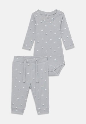 Costum de bebeluș gri deschis cu mâneci lungi și pantaloni, având un model cu bărci cu pânze. Fabricat dintr-un material ribbed moale, cu o croială ajustată.