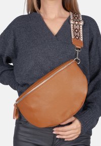 Sac croisé en cuir beige avec une surface texturée, doté d'une fermeture éclair argentée et d'une bandoulière à motifs, porté sur un pull sombre.