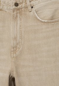 Beige spijkerbroek met voorzak, metalen knoopsluiting en zichtbare stiksels langs naden en zakranden.