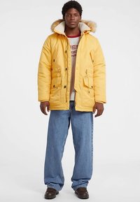 Geel geïsoleerd parka met een capuchon afgezet met bont, voorzien van grote voorzakken, een rits en knoopsluitingen. Draagt over een grijze trui en een blauwe spijkerbroek.