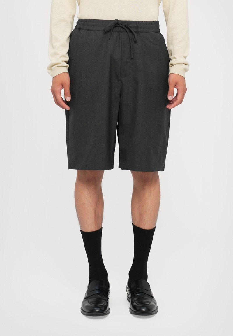 Graue, maßgeschneiderte Shorts mit einem elastischen Bund und Kordelzug. Glatte Textur, knielang, kombiniert mit schwarzen Socken und Slippern.