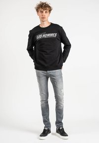 Sudadera negra con "LES HOMMES" impreso en blanco, combinada con jeans ligeros gris desgastados y zapatillas negras con suela blanca.