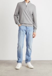 Man som bär ljusblå jeans, vita sneakers och en grå halvdragkedje-pullovertröja, står mot en enfärgad bakgrund.