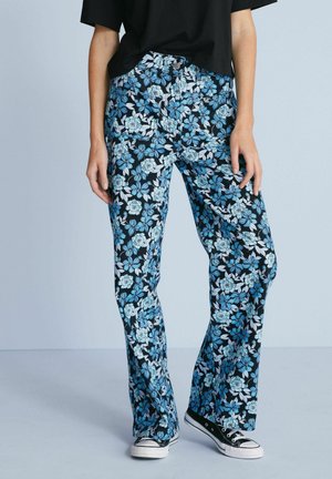 Persoon draagt blauw-witte broek met bloemenpatroon, zwart shirt en zwarte sneakers, staand tegen een lichte achtergrond.