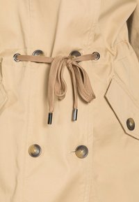 Chaqueta beige con cintura ajustable por cordón, que presenta ojales metálicos y dos botones decorativos. Textura suave con costuras uniformes.