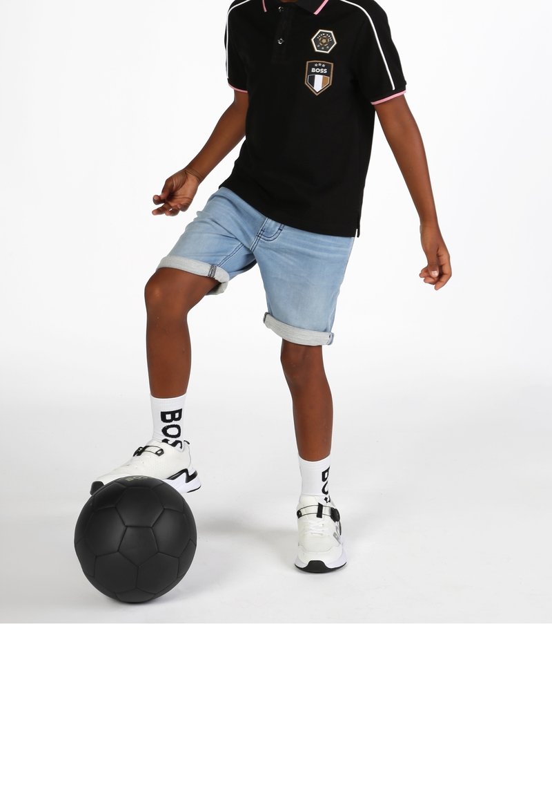 Enfant portant un polo noir, un short en jean et des baskets blanches contrôle un ballon de football noir avec le pied sur fond blanc.