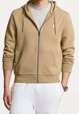 Homme portant un sweat à capuche marron clair avec fermeture éclair et poches, par-dessus un t-shirt gris clair et un pantalon blanc, montrant une montre-bracelet au poignet gauche.