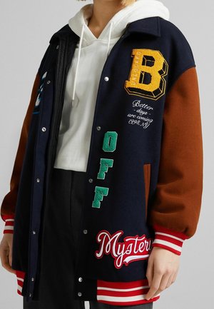 Osoba nosząca granatową i brązową kurtkę typu varsity z naszywkami, w tym dużą żółtą literą B oraz napisami „OFF” i „Mystery”. Pod spodem biała bluza z kapturem.