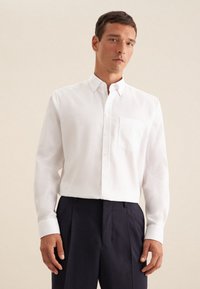 Camicia bianca con bottoni realizzata in tessuto liscio, con maniche lunghe, taschino sul petto e colletto classico, abbinata a pantaloni scuri.