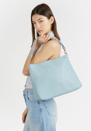 Giovane donna con capelli castani lisci tiene una grande borsa celeste con manico intrecciato sulla spalla, indossa una maglietta a righe e jeans chiari.