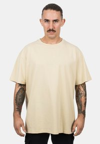 Beige cotton short-sleeve t-shirt with a round neckline. Relaxed fit and smooth texture. Visible black tattoos on arms.