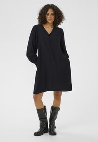 Robe bleu marine avec un décolleté en V, manches longues bouffantes et boutons sur le devant. Tissu lisse, coupe légèrement ample, associée à des bottes noires montantes jusqu'au genou.