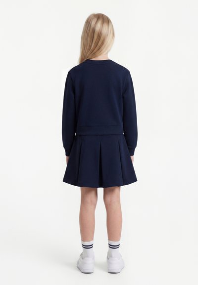 Enfant aux longs cheveux blonds vue de dos, portant un haut bleu marine à manches longues, une jupe plissée, des chaussettes blanches et des baskets blanches.