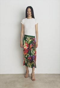 Bílý žebrovaný top s krátkým rukávem v kombinaci s dlouhou sukní s květinovým vzorem a zelenými akcenty, která má boční rozparek; model má na sobě béžové lodičky.