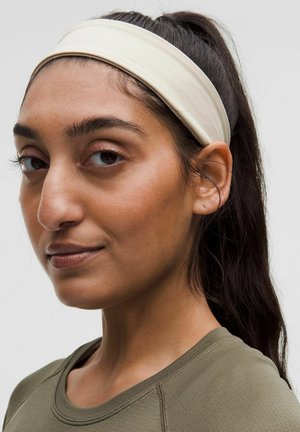 Mujer con cabello oscuro en una coleta, que lleva una diadema beige y una camiseta verde oliva, mirando ligeramente de costado contra un fondo simple.