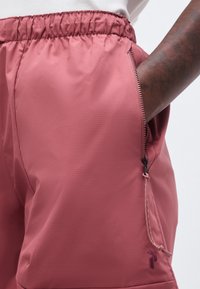 Το δεξί χέρι στην τσέπη με φερμουάρ σε ροζ παντελόνι με ελαστική μέση, εμφανίζοντας το τράβηγμα του φερμουάρ και μικρό κεντητό λογότυπο κοντά στο τελείωμα.