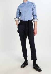 Světle modrá pruhovaná košile na knoflíky, tmavé elegantní kalhoty, černé boty a tmavý svetr uvázaný v pase. Čistý, ležérní vzhled.