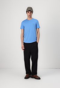 Hellblaues T-Shirt, Rundhals, Logodetail auf der Brust, lockere schwarze Hose und braune Schuhe. Hintergrund ist einfarbig weiß.