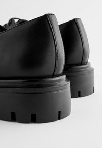 Scarpe in pelle nera con tacco alto e arrotondato, suola spessa e texturizzata, e cuciture visibili lungo i bordi del tacco.