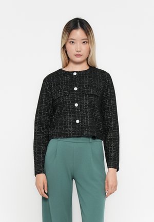 Veste noire courte avec un motif texturé et des boutons en argent, présentant un col rond et des manches amples, assortie à un pantalon vert.