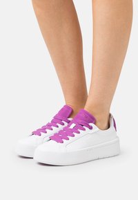 Weiße Sneakers mit lila Schnürsenkeln und gepolstertem Kragen. Verfügt über eine Gummisohle und ein strukturiertes Seitenpaneel. Markensymbol an der Ferse.