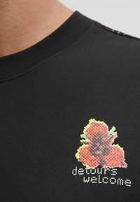 Tricou negru din bumbac cu decolteu rotund. Prezintă un grafic colorat pixelat al unei flori și textul "devierile sunt binevenite" în alb dedesubt.