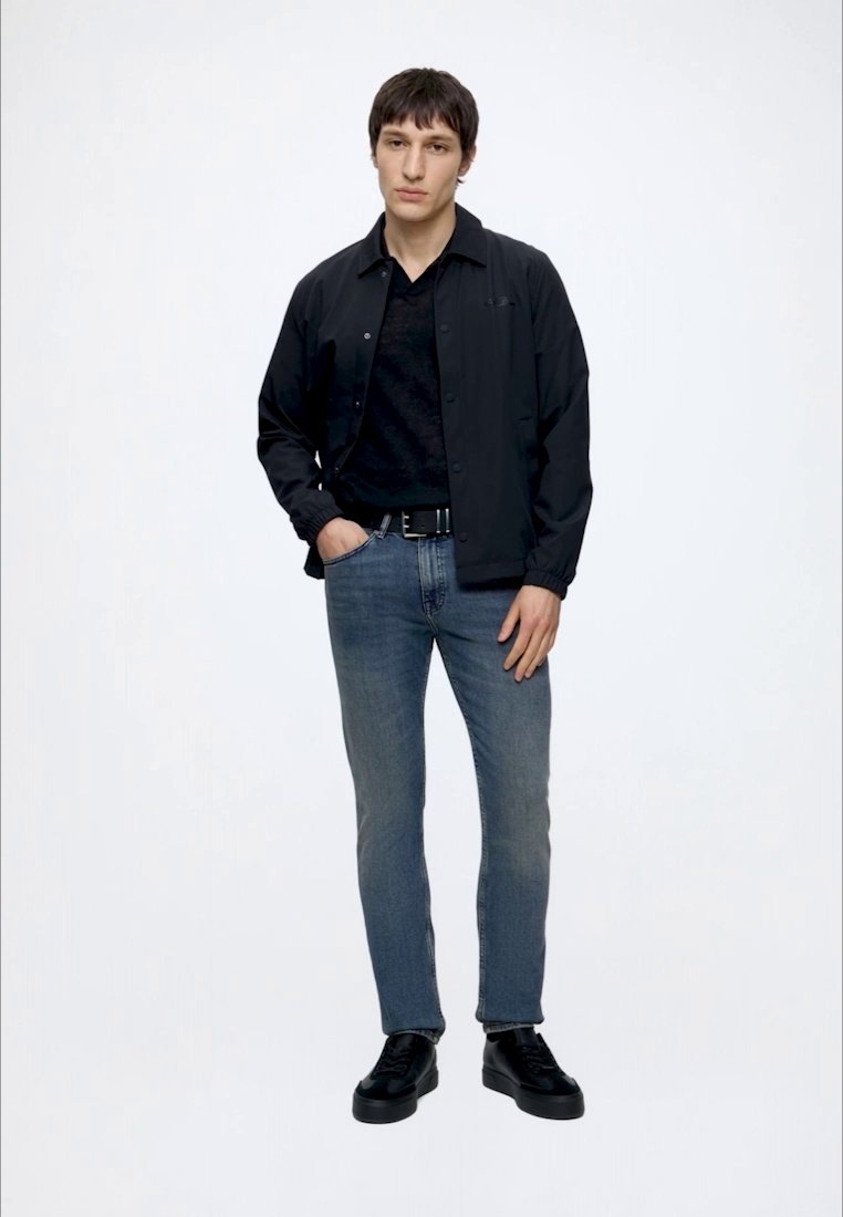 Jeune homme debout portant une veste noire, une chemise noire, un jean bleu, une ceinture noire et des chaussures noires sur un fond blanc uni.