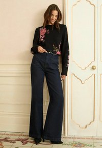 Melnā džemperis ar ziedu izšuvumiem, augstās jostas vietas tumšas džinsu bikses ar plašu kāju, un melnas augstpapēžu kurpes, pozējot uz gaiša fona.