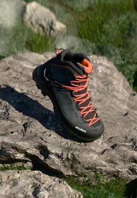 Black hiking boot with orange laces and trim, rubber sole, textured material, and high ankle support, displayed on a rocky surface.