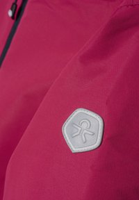 Fuchsia-Jacke aus glattem, wasserabweisendem Material mit einem strukturierten grauen Logo-Patch in Form eines Sechsecks am Ärmel.