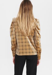 Blusa giallo dorato a scacchi con maniche a sbuffo, texture a coste e polsini con bottoni. Indossata con pantaloni neri per creare un contrasto.