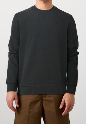 Mann trägt ein schlichtes schwarzes Sweatshirt mit langen Ärmeln und braune Hose, steht mit entspannt an den Seiten hängenden Armen vor einem einfarbigen Hintergrund.