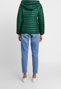 Grøn isoleret jakke med hætte, der har lodrette syninger og lynlås, stylet med lyseblå jeans og hvide sneakers.