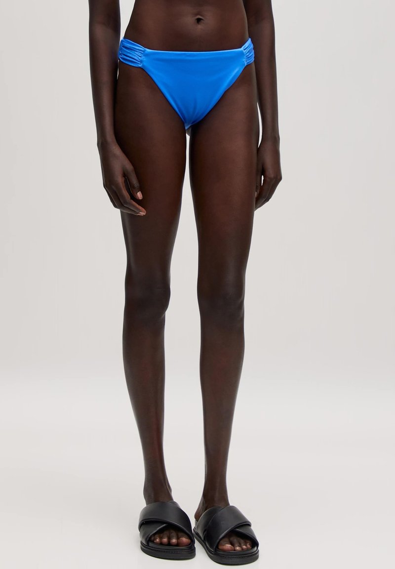 Blaue Bikiniunterteile mit gerafften Seitendetails, aus glattem Material gefertigt. Kombiniert mit schwarzen Slipper-Sandalen mit einem Kreuzgurt-Design.