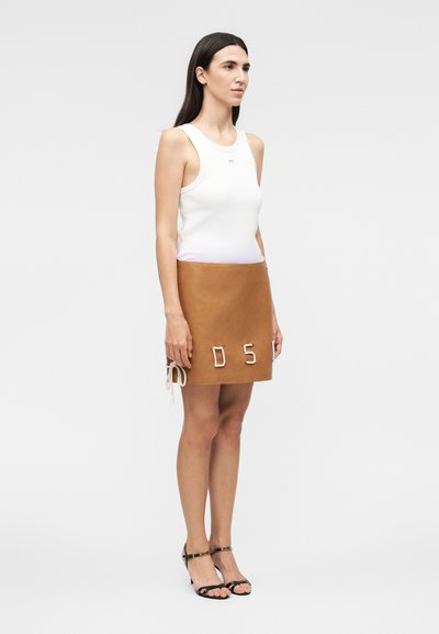 Γυναίκα με μακριά σκούρα μαλλιά, που φοράει λευκό αμάνικο τοπ, καφέ μίνι φούστα με λεπτομέρειες από σχοινί και μαύρα πέδιλα με τακούνι, που στέκεται πλαγίως.