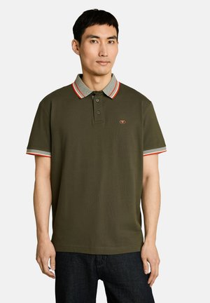 Homme portant un polo vert olive avec un liseré gris et rouge au col et aux manches, associé à un jean foncé, debout devant un fond uni.