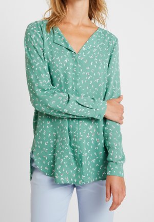 Femme portant un chemisier vert à manches longues avec des motifs de feuilles blanches et roses, associé à un pantalon bleu clair, bras croisés sur la poitrine.