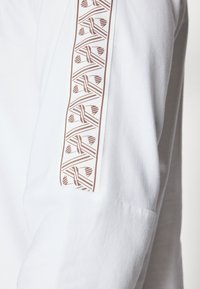 Witte lange mouw top met een bruine geometrische patroon op de mouwen, een gladde textuur en een getailleerd ontwerp.