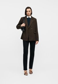 Νεαρή γυναίκα με μαύρο σακάκι, λευκό πουκάμισο με γιακά, μαύρο παντελόνι, ψηλοτάκουνα με λουράκια, κρατώντας μαύρη τσάντα στον ώμο, στέκεται μπροστά σε απλό φόντο.