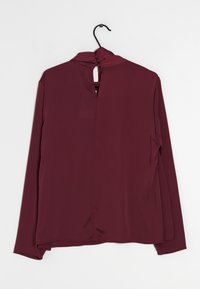 Blusa bordeaux a maniche lunghe con scollatura drappeggiata, apertura a chiave sul retro e superficie liscia. Appesa a un gancio nero.